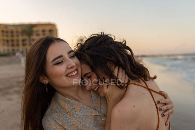 Молоді веселі подружки обіймаються один з одним, стоячи на піщаному пляжі біля махаючого моря на заході сонця — стокове фото