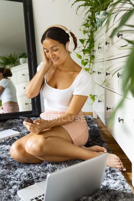 Cheerful ethnic female with cellphone listening to song from wireless headphones while sitting on carpet against mirror at home — Stock Photo