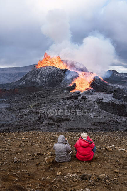 Vista posteriore di viaggiatori irriconoscibili che ammirano Fagradalsfjall con fuoco e lava mentre scattano foto e si siedono sul monte in Islanda — Foto stock