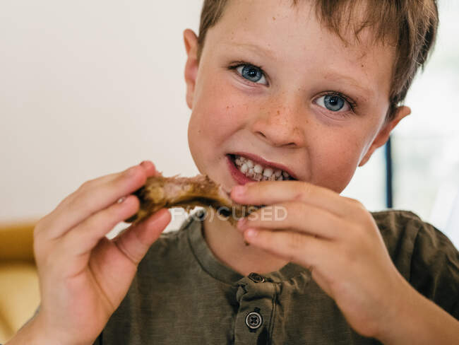 Primo piano di adorabile bambino mangiare costolette di maiale appetitoso durante il pranzo a casa e guardando la fotocamera — Foto stock