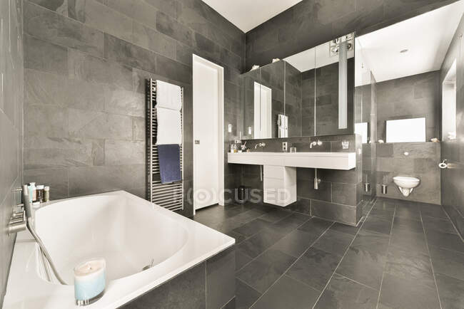 Intérieur de salle de bain spacieuse avec des murs en carrelage gris et sol et baignoire blanche et lavabos — Photo de stock