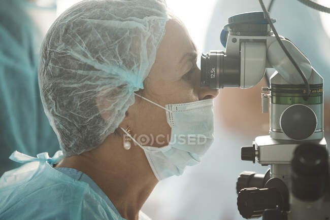 Орієнтована доросла жінка-лікар у стерильній масці та декоративній медичній кепці, яка дивиться через хірургічний мікроскоп проти колеги по сільськогосподарській галузі в лікарні — стокове фото