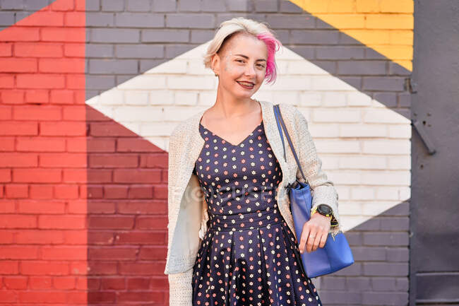 Happy fêmea alternativa no vestido da moda e com cabelo curto tingido de pé contra a parede de tijolo colorido na rua e olhando para a câmera — Fotografia de Stock