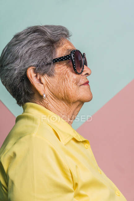 Вид збоку на щасливу сучасну літню жінку з сірим волоссям і в модних сонцезахисних окулярах на рожевому фоні в студії і дивиться в сторону — стокове фото