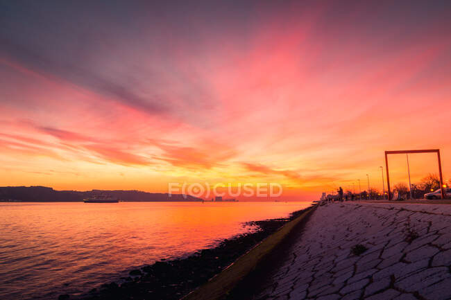 Pendio di pietra di argine situato vicino increspatura fiume Tago contro cielo nuvoloso tramonto rosso a Lisbona, Portogallo — Foto stock
