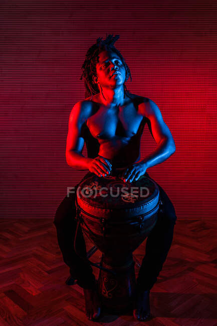 Dreamy black male musician with naked torso playing African drum in studio with red and blue neon lights — Stock Photo