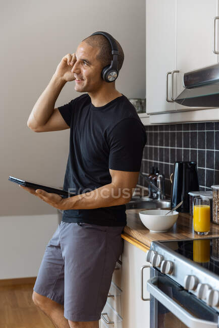 Hombre contento disfrutando de la música en los auriculares mientras está de pie con la tableta en la cocina durante el desayuno por la mañana y mirando hacia otro lado - foto de stock