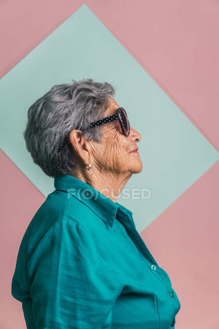 Vista lateral da mulher moderna feliz envelhecida com cabelos grisalhos e em óculos de sol na moda em fundo rosa e azul em estúdio e olhando para longe — Fotografia de Stock
