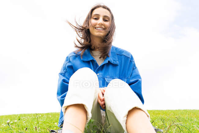 Bajo ángulo de alegre hembra sentada en la colina en la naturaleza en un día ventoso mientras mira hacia otro lado - foto de stock