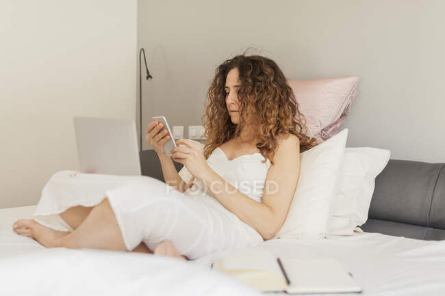 Vista lateral de la mujer de negocios con el pelo rizado sentado en la cama y trabajando con su computadora portátil y teléfono inteligente - foto de stock