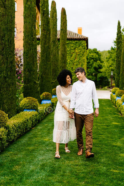 Cheerful multiracial couple holding hands and walking along green path in alley in garden while looking at each other — Stock Photo