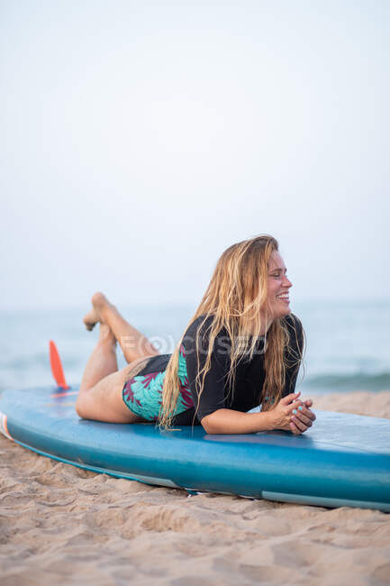 Vista lateral del surfista femenino positivo en traje de baño acostado en el paddleboard en la orilla arenosa contra el mar y mirando hacia otro lado - foto de stock