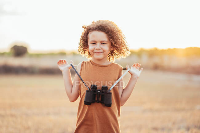 Criança étnica bonito com cabelos cacheados e binóculos em pé no campo seco no verão e olhando para baixo — Fotografia de Stock