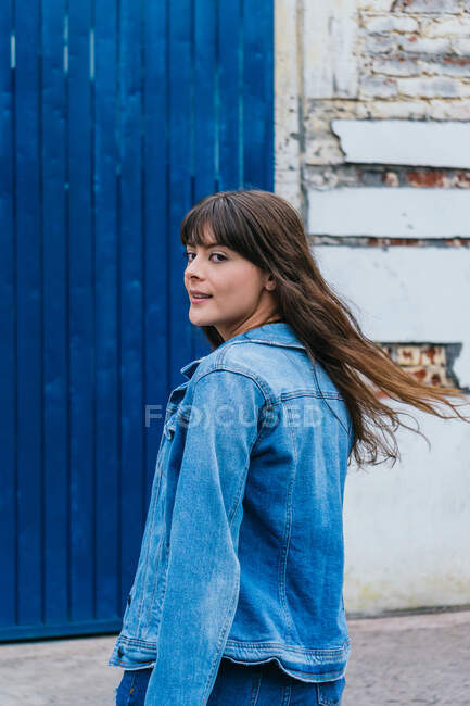 Femme tranquille avec de longs cheveux bruns volants et veste en denim regardant la caméra sur fond bleu en ville — Photo de stock