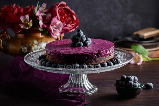 Delicioso bolo de mousse de mirtilo com creme roxo decorado com bagas frescas servidas em suporte de vidro na mesa escura com flores — Fotografia de Stock