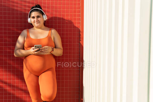 Cheerful ethnic female athlete with curvy body and cellphone listening to song from headphones while looking at camera against tiled wall — Stock Photo
