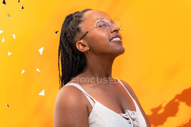 Side view of dreamy African American female with braided hairstyle and in glasses standing with closed eyes on yellow background in Barcelona and enjoying summer — Stock Photo