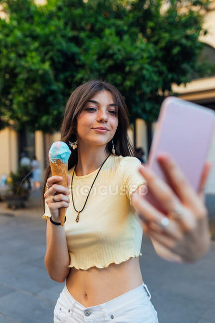 Дружелюбная женщина с вкусным мороженым в вафельном конусе делает автопортрет на сотовом телефоне на городской тротуаре — стоковое фото