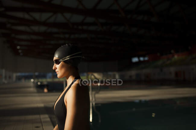 Молода красива жінка, що сидить на краю критого басейну з чорним купальником, дивиться на камеру — стокове фото