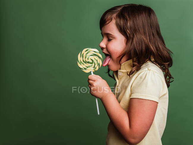 Вид збоку смішний дев'ятнадцять дітей лизати солодкий вихровий льодяник на зеленому фоні в студії — стокове фото