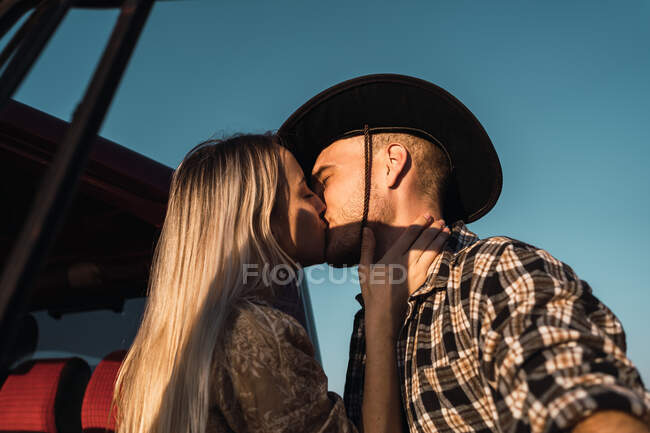 Desde abajo vista lateral de la joven mujer cariñosa besar hombre en sombrero de vaquero tiernamente cerca del coche en el fondo del cielo azul por la noche - foto de stock