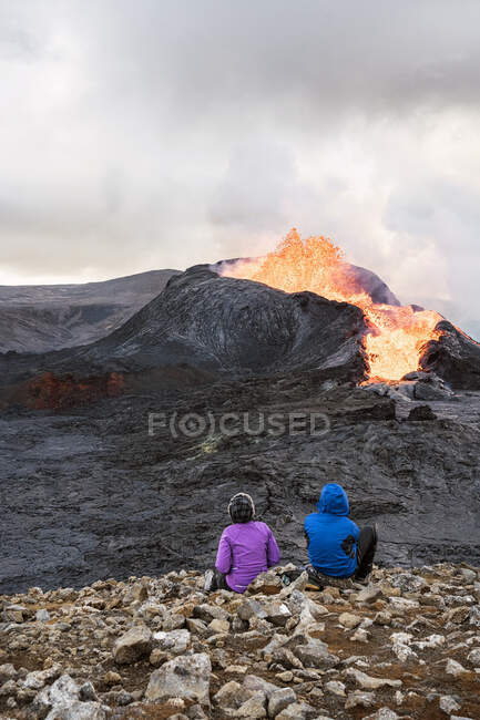 Back view of unrecognizable travelers admiring Fagradalsfjall with fire and lava while sitting on mount in Iceland — Stock Photo