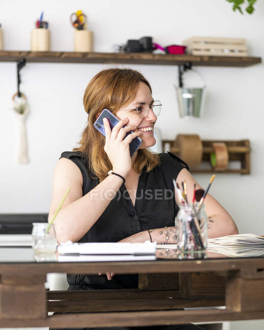 Content female artist sitting at table with paintings and speaking on mobile phone while discussing project in workspace — Stock Photo