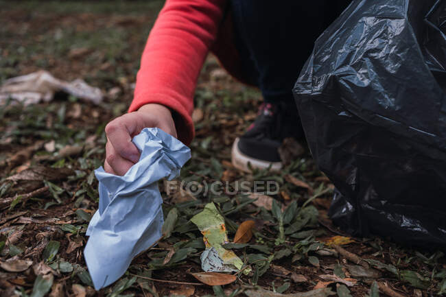 Cropped unrecognizable volunteer with plastic bags picking rubbish from terrain against trees in summer woods in daylight — Stock Photo