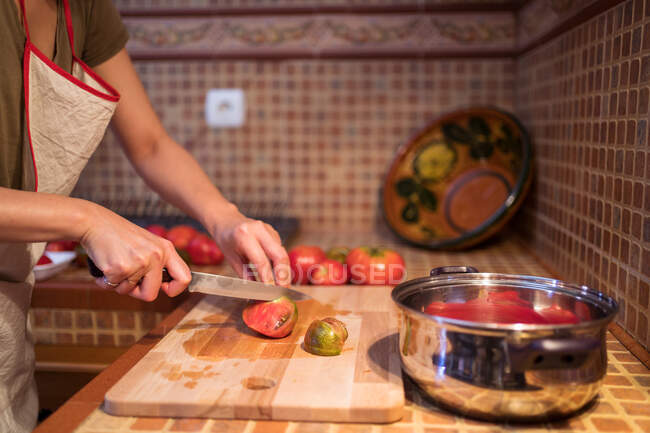 Vue latérale d'une femme ethnique dans un tablier découpant des tomates mûres sur une planche à découper pendant la cuisson du déjeuner dans la cuisine à la maison — Photo de stock