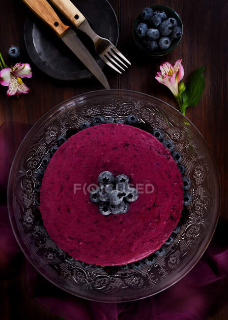 Deliziosa torta di mousse di mirtilli con crema viola decorata con bacche fresche servite su supporto di vetro su tavolo scuro con fiori — Foto stock