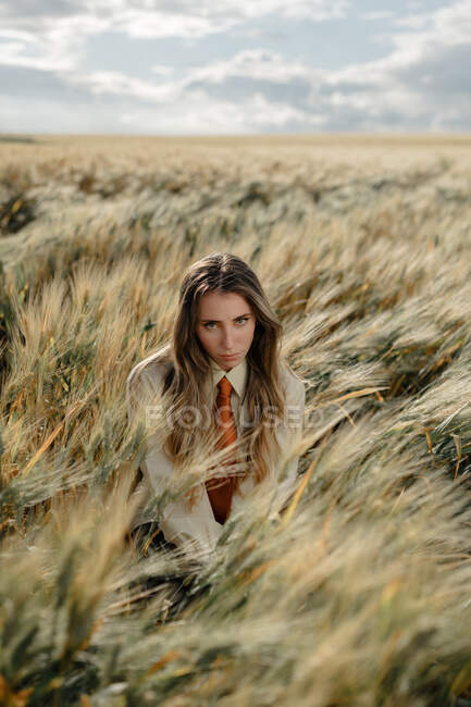 Young female with wavy hair looking at camera in countryside field under cloudy sky on blurred background — Stock Photo