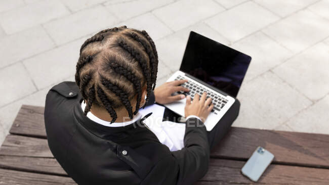 D'en haut de freelance masculin ethnique méconnaissable avec coiffure tressée assis sur le banc et tapant sur le clavier de l'ordinateur portable tout en travaillant sur le projet à distance sur la rue urbaine — Photo de stock