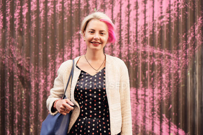 Чарівна альтернативна жінка з фарбованим волоссям і в модному одязі, що стоїть на тлі фіолетової стіни в місті і дивиться на камеру — стокове фото