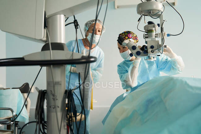 Doctora atenta en uniforme estéril contra su compañero de trabajo que mira hacia otro lado mientras se prepara para la cirugía en el hospital con microscopio - foto de stock