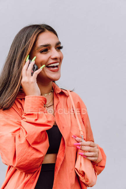 Mujer alegre con uñas largas y brillantes de pie cerca de la pared blanca y hablando en el teléfono celular mientras se ríe felizmente - foto de stock