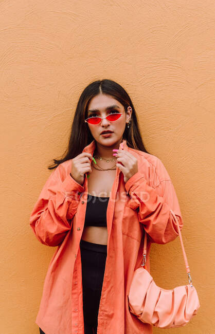 Уверенная молодая женщина тысячелетия в модном наряде и в современных солнцезащитных очках, стоящая с мешком у оранжевой стены — стоковое фото