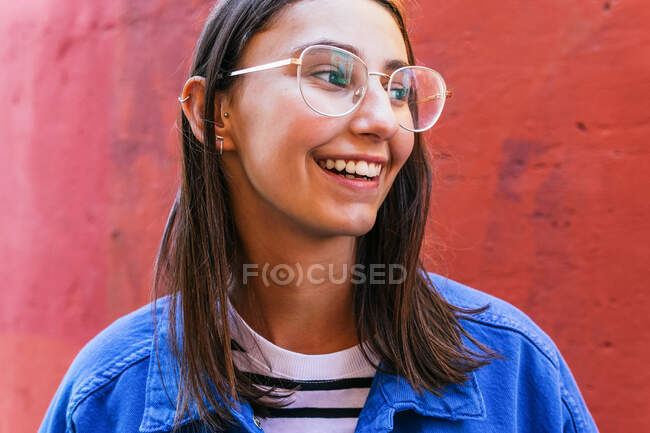 Femme positive en tenue élégante regardant loin sur fond coloré de mur de construction par jour ensoleillé dans la rue de la ville — Photo de stock