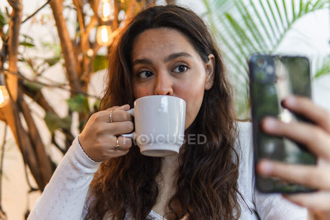 Jovem latino-americana fêmea tirando selfie no celular enquanto bebe café no café com plantas verdes no fundo — Fotografia de Stock