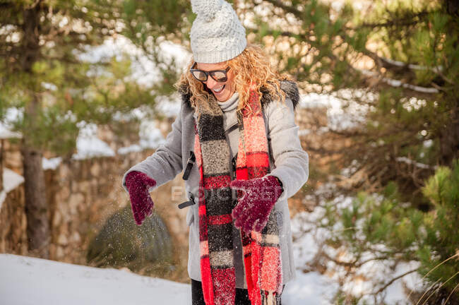 Delighted female in warm clothes and sunglasses tossing snow and having fun in winter woods — Stock Photo