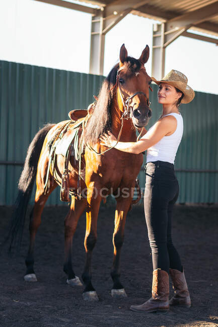 Side view of content young female caressing muzzle of mare on sandy land under roof in countryside on sunny day — Stock Photo