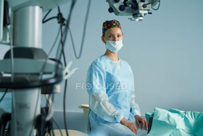 Femme médecin adulte en uniforme chirurgical et masque stérile regardant la caméra alors qu'elle était assise à la clinique — Photo de stock