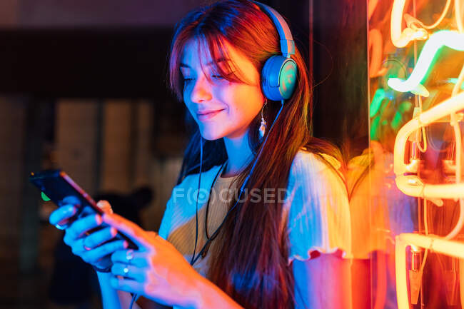 Fröhliche junge Frau mit Handy hört Lied aus Kopfhörern gegen bunte Neonlichter in der Abendstadt — Stockfoto