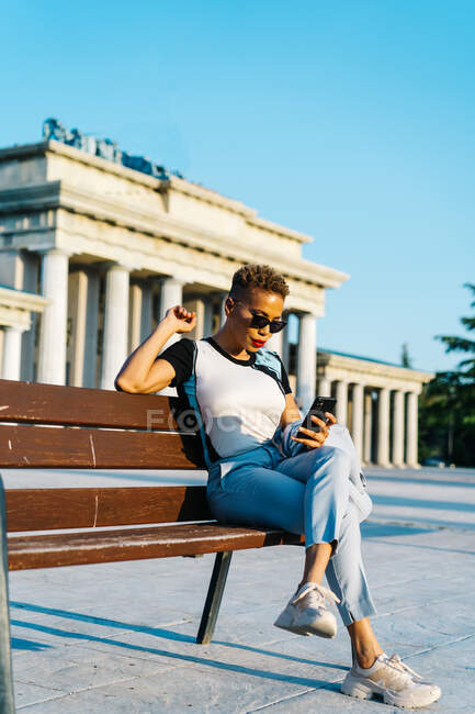 Feminino étnico na moda em óculos de sol e corte de cabelo moderno usando smartphone enquanto sentado no banco na cidade — Fotografia de Stock
