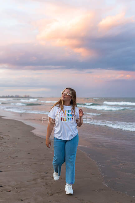 Positive junge Frau in trendiger Sonnenbrille und stylischem Outfit, die an einem Sommerabend am Meer gegen das Meer läuft — Stockfoto