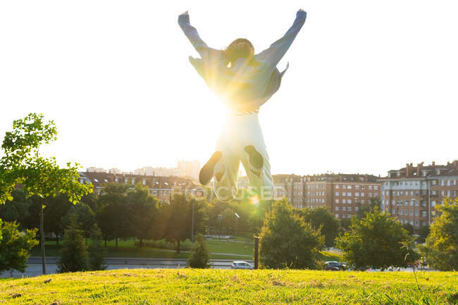 Back view of unrecognizable carefree female with raised arms in moment of jumping above hill against city in back lit — Stock Photo