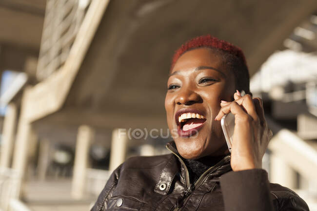 Seitenansicht der schönen schwarzen Afro-Frau, die mit ihrem Smartphone spricht, während sie an einem sonnigen Tag mit verschwommenem Hintergrund lächelt — Stockfoto