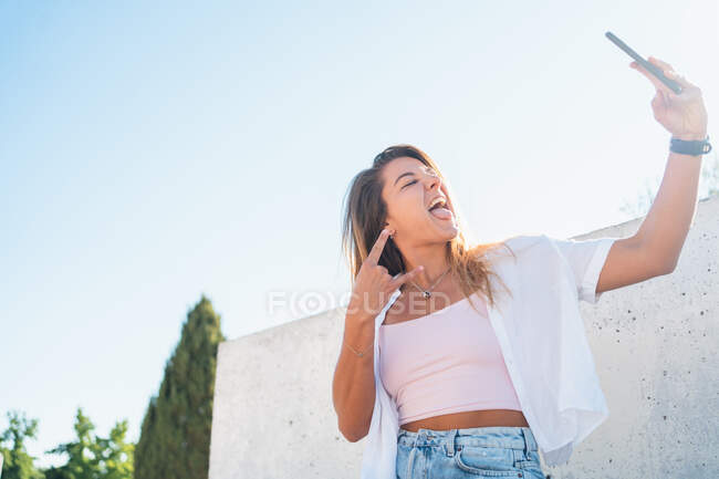 Знизу позитивно весела молода жінка стирчить язиком і робить рок-н-рол жест, приймаючи самостріл на мобільний телефон в сонячний день влітку в місті — стокове фото