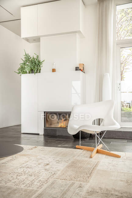 Chaise blanche placée sur le tapis près de la cheminée avec des plantes décoratives dans la maison lumineuse moderne — Photo de stock