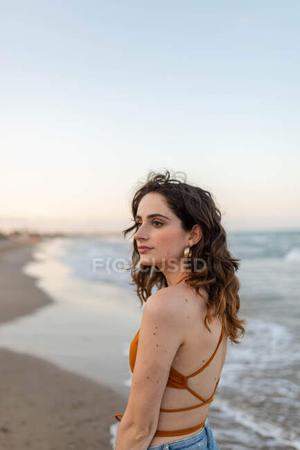 Sonhador jovem fêmea com cabelos longos olhando para longe, enquanto em pé na praia de areia perto do mar ondulando — Fotografia de Stock