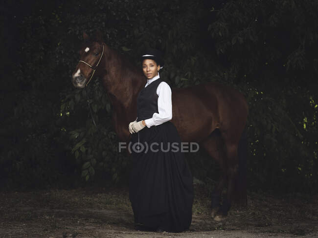 Confident African American adult lady in elegant clothes and hat standing with brown horse while looking at camera near trees in daytime — Stock Photo